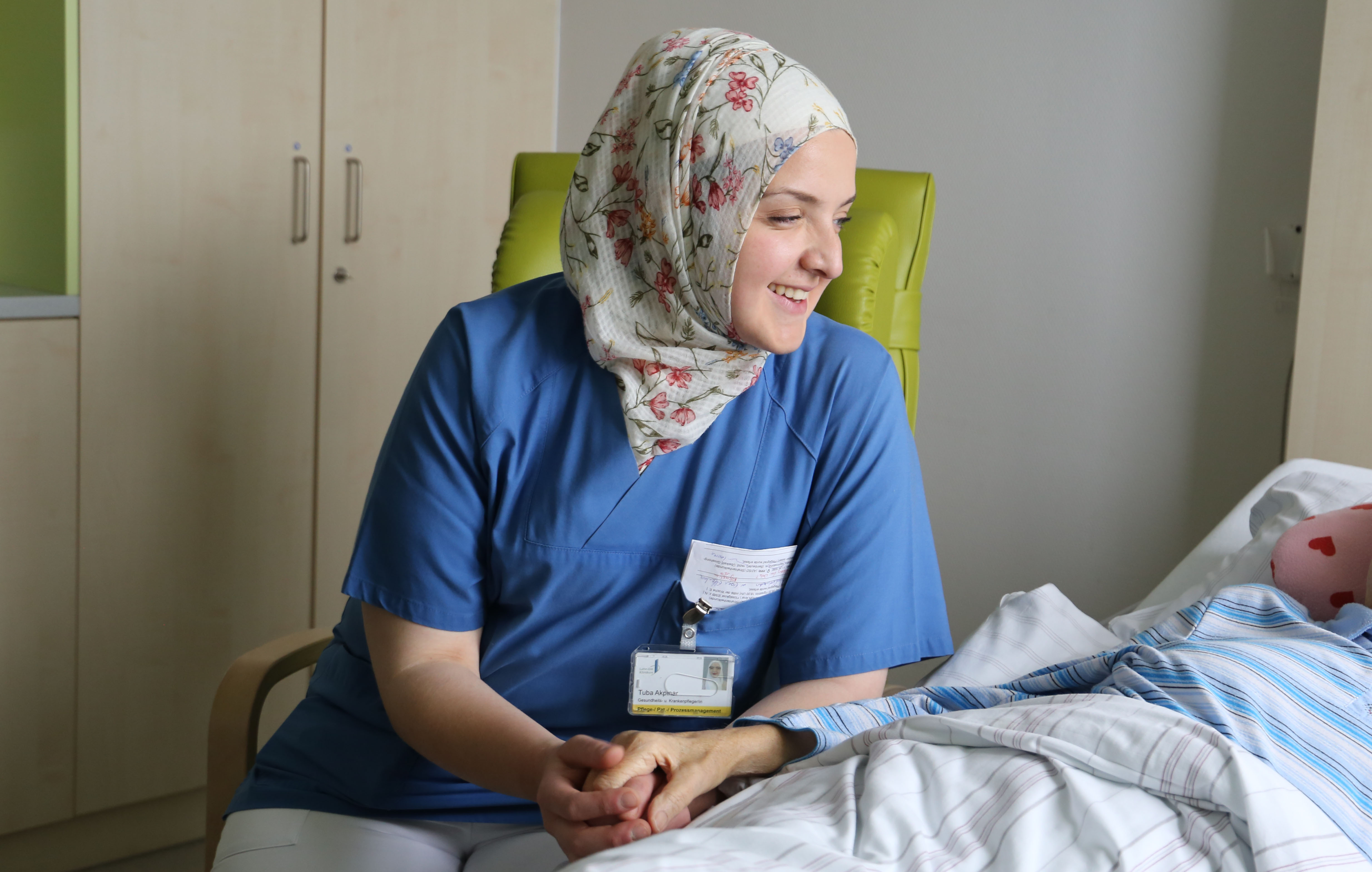 Pflegekraft mit Migrationshintergrund sitzt am Bett eines Patienten und hält die Hand. 